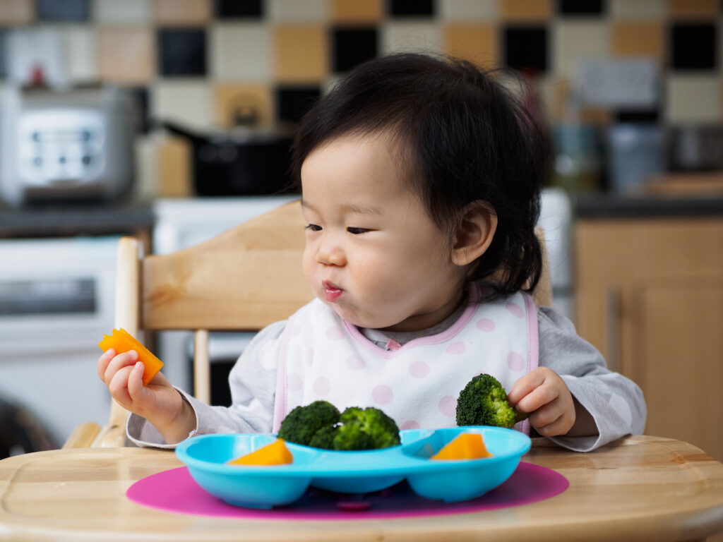 Baby at table eating plate of butternut squash and broccoli
